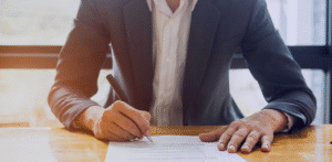 Close-up of someone signing paperwork with a pen on a wooden desk
