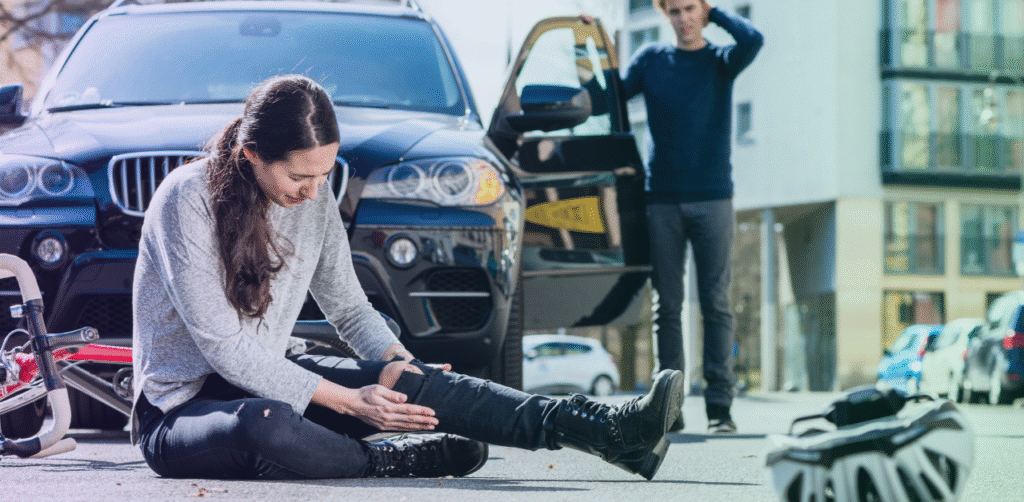 Injured cyclist sits on the road holding her leg beside a fallen bicycle and stopped SUV