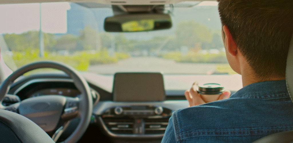 Person seated in the front passenger seat of a car, holding a takeaway coffee cup while looking through the windshield