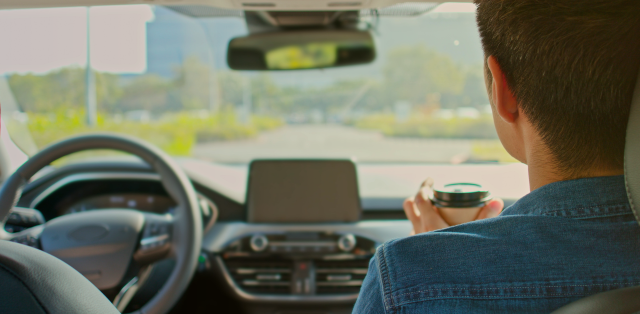 Person seated in the front passenger seat of a car, holding a takeaway coffee cup while looking through the windshield