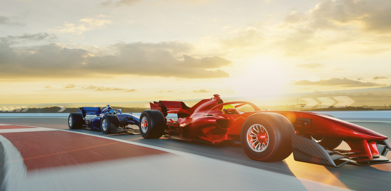 Two open-wheel race cars speed side by side around a racetrack at sunset, with a red car in the foreground and a blue car close behind