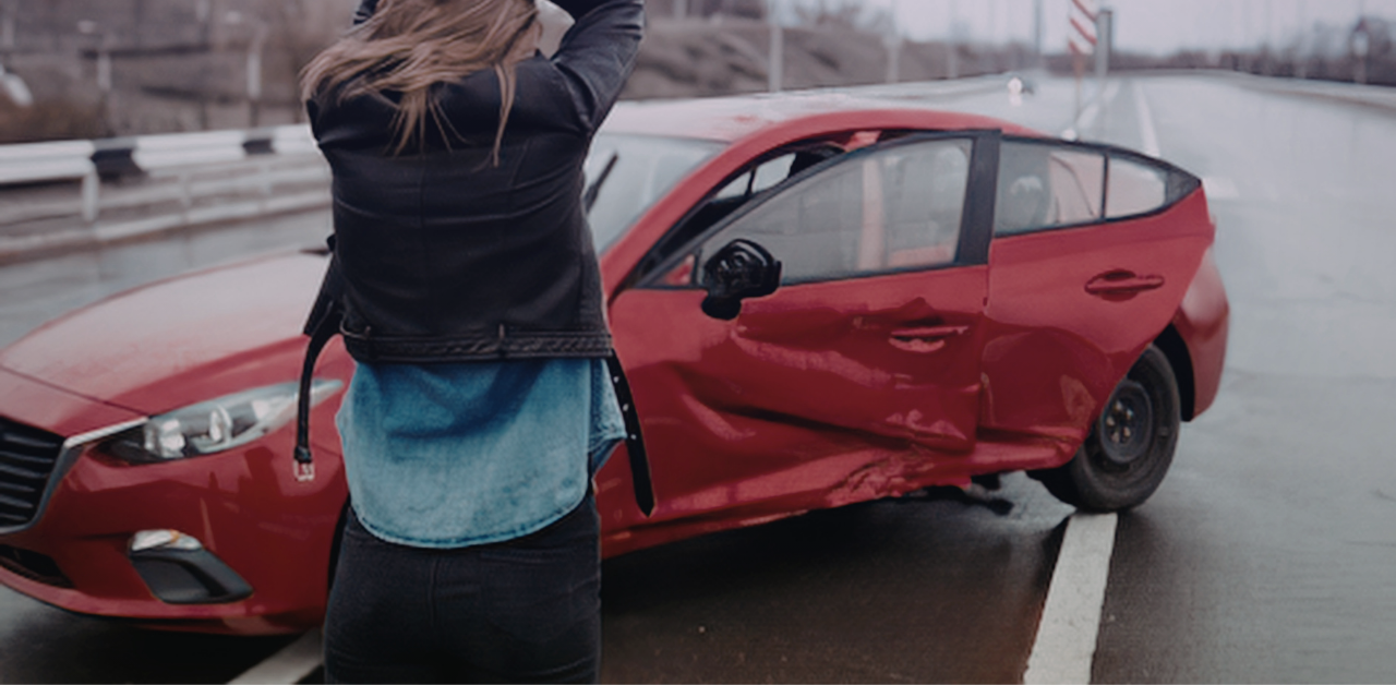 Woman stands beside a red car with severe side damage after a collision on a wet roadway