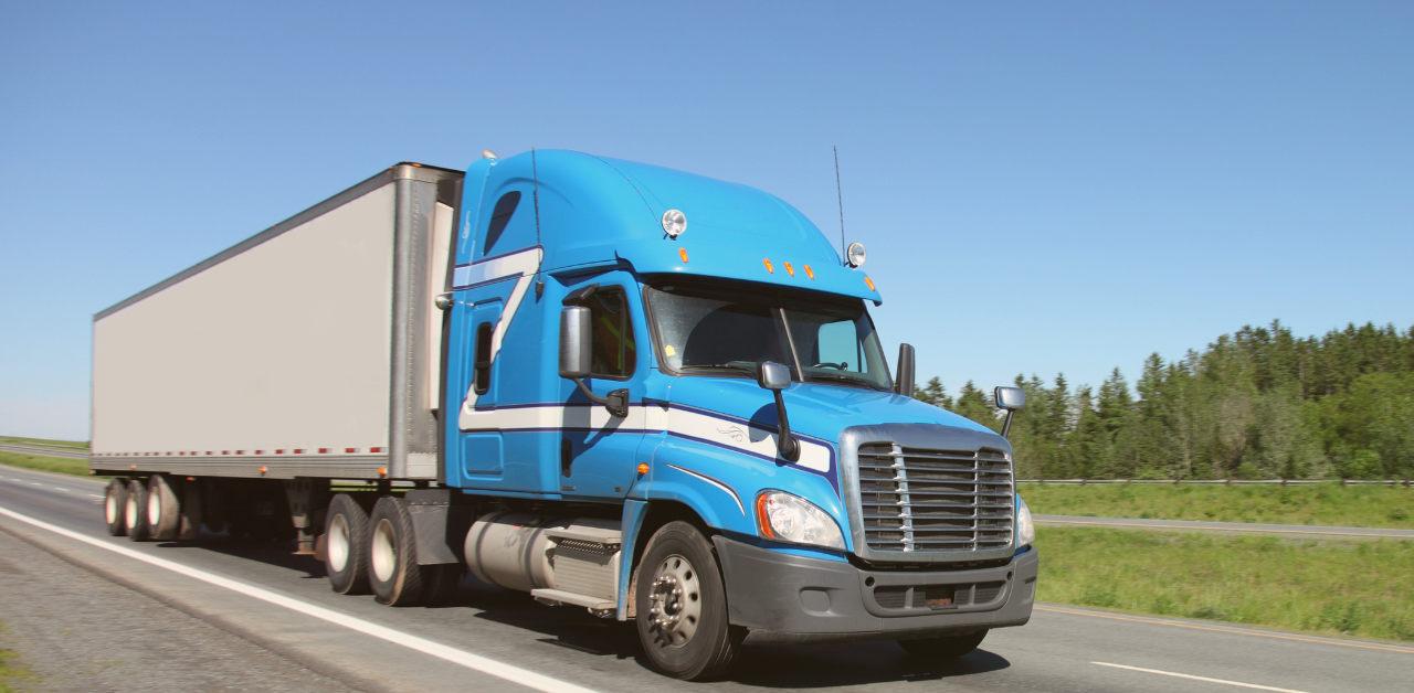 A blue semi-truck hauling a white trailer travels along a highway on a clear day