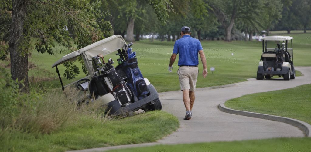 A golfer walks toward a golf cart that has veered off the paved path and tilted into a grassy ditch