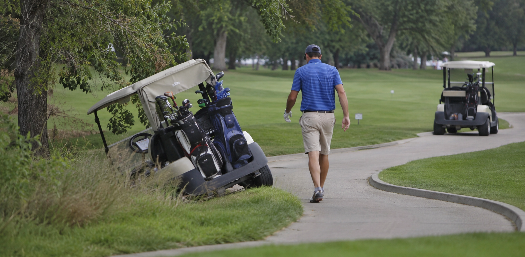 A golfer walks toward a golf cart that has veered off the paved path and tilted into a grassy ditch