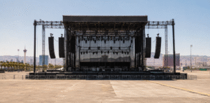 A large outdoor concert stage with hanging speakers and lighting equipment stands in an empty event area