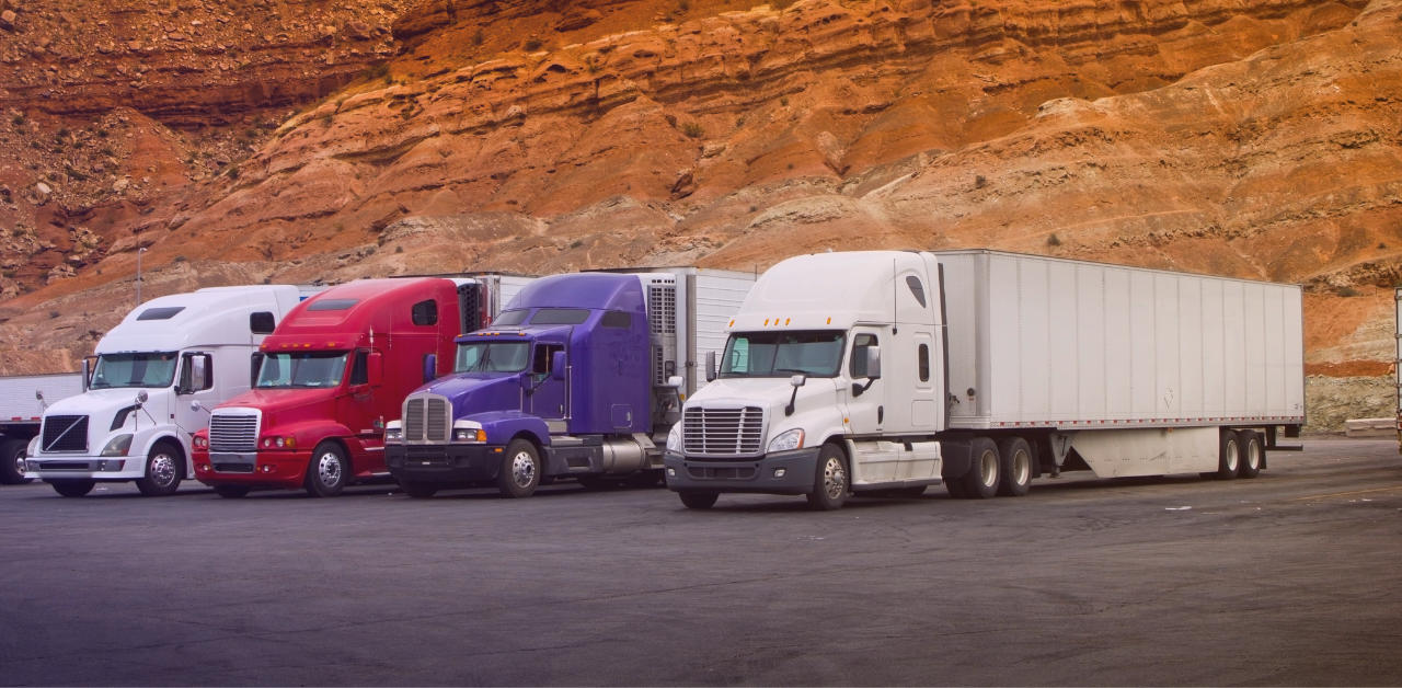 A row of tractor-trailers rests in a paved lot surrounded by steep, rust-colored desert rock formations