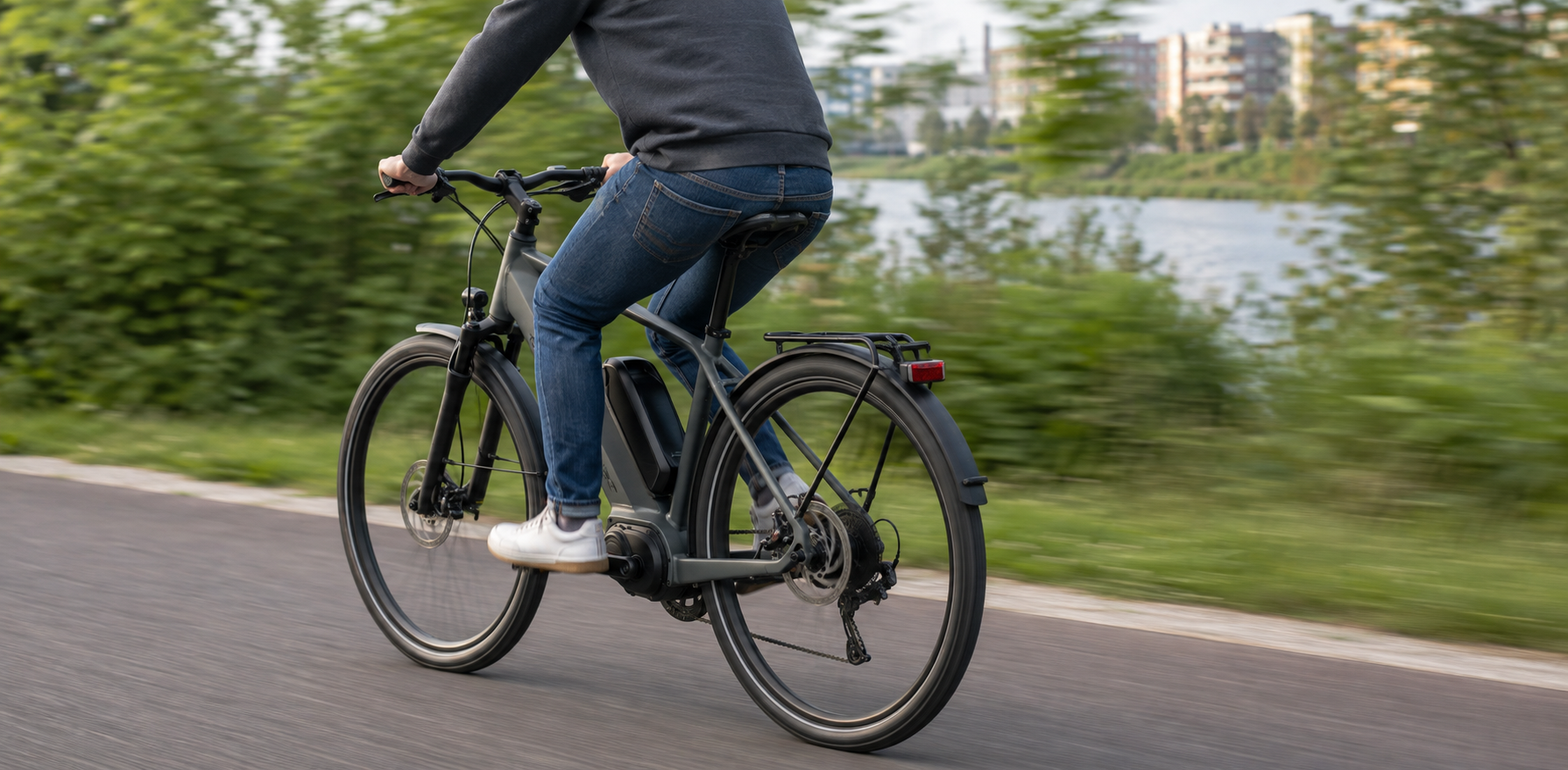 Cyclist rides an electric bicycle along a paved path beside greenery and water, with buildings visible in the distance