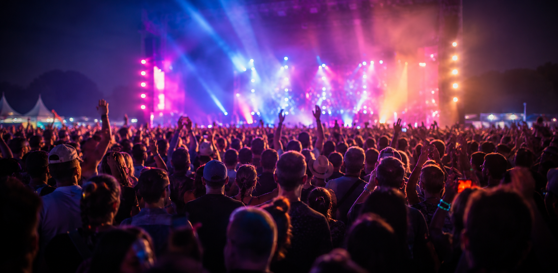 Hundreds of concertgoers raise their hands toward a brightly lit outdoor stage filled with blue, pink, and orange lights