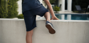 Rear view of a child climbing over a low wall toward an outdoor pool, illustrating a potential pool safety hazard