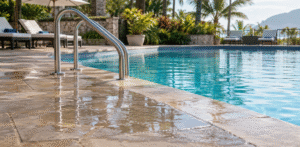 Wet stone-tiled swimming pool deck with puddles of water surrounding stainless steel handrails beside a clear blue pool